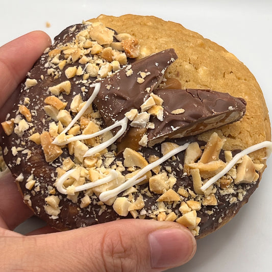 Close-up of The Dulce de Leche Bliss Cookie topped with Snickers, peanuts, and white chocolate drizzle